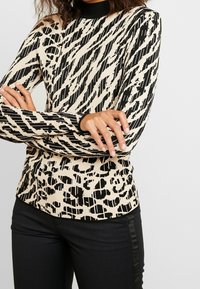 Top aderente a maniche lunghe in un motivo geometrico beige e nero, con un colletto alto a costine e un tessuto testurizzato. Abbinato a pantaloni neri.
