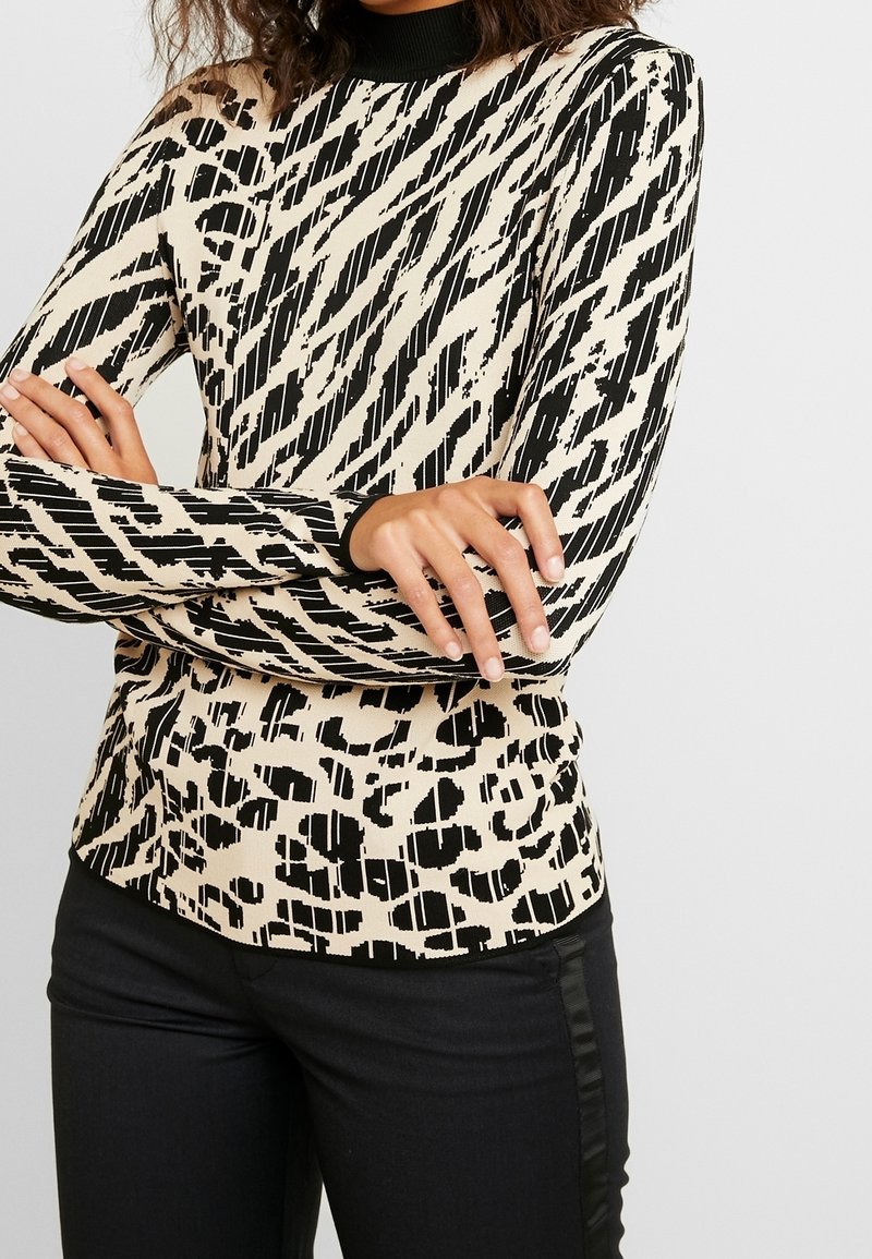 Top aderente a maniche lunghe in un motivo geometrico beige e nero, con un colletto alto a costine e un tessuto testurizzato. Abbinato a pantaloni neri.