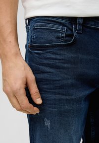 Ciemnoniebieskie dżinsy mają dopasowany krój, z minimalnym przetarciem na materiale i dłonią opartą na udzie.