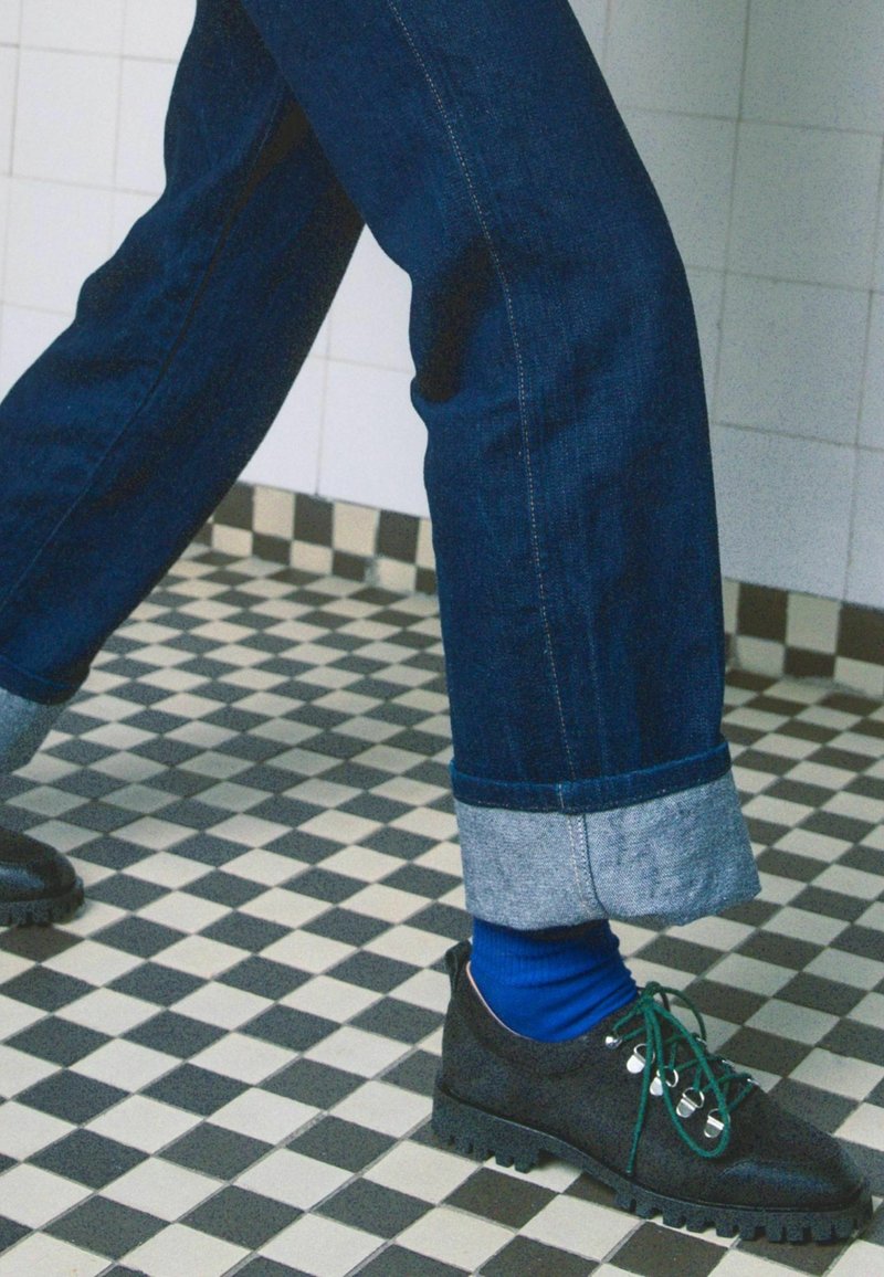 Scarpe nere con lacci verdi, suole in gomma grigia, abbinate a jeans in denim scuro con un risvolto azzurro chiaro, su un pavimento a quadri.