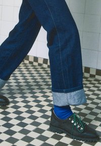Tmavě modré džíny z denimové látky s ohrnutými nohavicemi, v kombinaci s černými botami a modrými ponožkami. Boty mají zelené tkaničky a texturovanou gumovou podrážku.