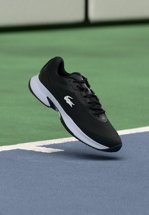 Black athletic shoe with a textured upper, featuring a white logo, lace detailing, and a contrast white and black sole, set against a court surface.