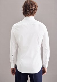 Camicia bianca a maniche lunghe realizzata in tessuto liscio, con polso abbottonato e orlo leggermente curvo, vista di spalle su uno sfondo neutro.