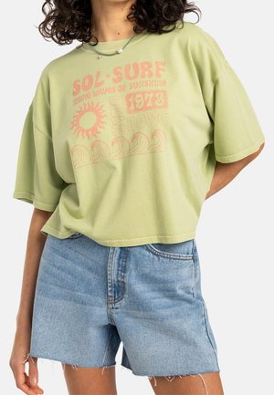 Kvinde iført lysegrøn oversized t-shirt med lyserøde surf-grafikker og lyseblå denimshorts med frynset kant, stående med den ene hånd bag ryggen.