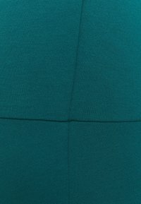 Teksturwana tkanina w kolorze niebiesko-zielonym z prążkowanym wzorem, z pionowymi szwami i geometrycznym designem. Materiał wydaje się elastyczny i gładki.