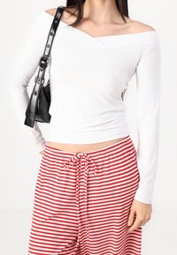 Bílá halenka s dlouhým rukávem a off-shoulder designem, doplněná červenými a bílými pruhovanými kalhotami na stažení. Černá kabelka s lesklým povrchem.