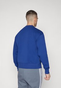 Felpa blu con collo tondo, polsini e orlo a costine. Tessuto morbido, colore uniforme, presenta un'etichetta sul lato sinistro. Abbinata a pantaloni grigi.