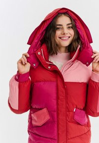 Usmívající se žena upravuje kapuci na barevném růžovém a červeném péřovém kabátu na jednoduchém bílém pozadí.