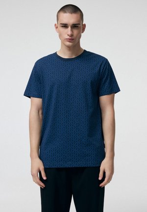 Młody mężczyzna w niebieskiej koszulce z krótkim rękawem w wzory i czarnych spodniach stoi na jasnym, jednolitym tle.