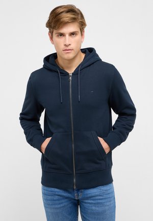 Young man wearing a navy blue zip-up hoodie with hands in front pockets and light blue jeans, facing forward against a white background.