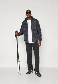 Zwarte sportjas met patroon en elastische manchetten, gecombineerd met zwarte broek. Accessoires zijn onder andere een wandelstok en donkere zonnebril.