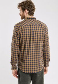 Homme aux cheveux bouclés portant une chemise à manches longues à carreaux marron et bleu marine, faisant face à un fond uni.