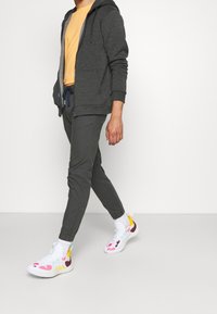 Grijze zip-up hoodie, gele t-shirt, grijze broek en kleurrijke sneakers met patronen. Sneakers hebben roze en paarse accenten.