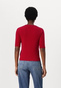 Roter gerippter Pullover mit kurzen Ärmeln und rundem Ausschnitt, kombiniert mit hochtaillierten blauen Jeans, die ein einfaches Design für die Gesäßtaschen aufweisen.