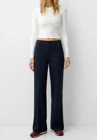 Pantaloni a gamba larga blu navy realizzati in tessuto liscio, con chiusura frontale a bottone e due tasche laterali, abbinati a una maglietta aderente bianca.