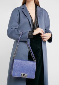 Borsa a spalla in pelle viola con forma rettangolare, dotata di una tracolla in catena argentata e di una chiusura in metallo. Texture liscia con una sottile lucentezza.
