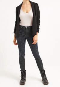 Schwarze gestrickte Strickjacke über einem hellgrauen Tanktop, kombiniert mit dunklen Jeans im Skinny-Fit und schwarzen Schnürstiefeln.