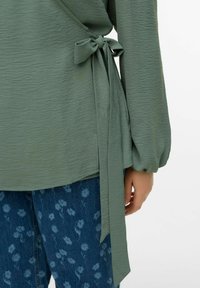 Grønn omslagstopp i teksturert stoff, med lange ermer og knyttedetalj i midjen, kombinert med blå jeans med blomsterprint.