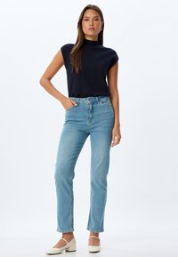 Top sin mangas en navy combinado con jeans de tiro alto en azul claro, con corte recto y un ligero desvanecimiento; la modelo lleva puestos zapatos de tacón bloque blancos.