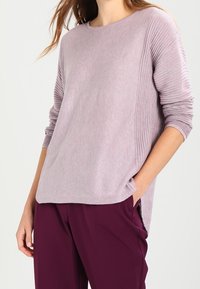 Suéter morado claro acanalado con escote redondo y mangas largas, con un ajuste holgado y aberturas laterales, combinado con pantalones morados oscuros.