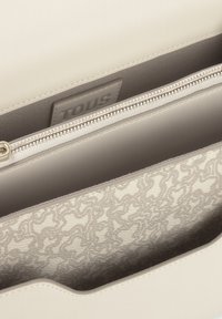 Bolso de mano de cuero beige claro con un exterior suave, cierre de cremallera y un forro interior texturizado que presenta un sutil patrón ondulado. Etiqueta de la marca visible.
