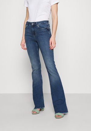 Femme portant un jean bootcut taille haute bleu foncé, un t-shirt blanc à manches courtes et des sandales à talons vertes tressées à bout ouvert, debout.