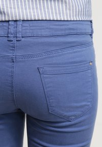 Jean en denim bleu au design ajusté, doté d'une poche arrière unique et de détails de couture subtils, tissu texturé et finition lisse.