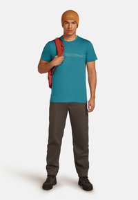 Muž v hnědém čepici a tyrkysové košili, který drží červenou bundu přes rameno, oblečený v hnědých kalhotách a černých turistických botách.