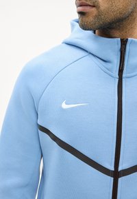 Homme portant un sweat à capuche zippé Nike bleu clair avec une bande diagonale noire et un logo swoosh blanc, cadré pour montrer le bas du visage et le haut du torse.