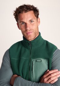 Man with short curly brown hair and blue eyes wearing green fleece vest over gray long-sleeve shirt, arms crossed, neutral background.