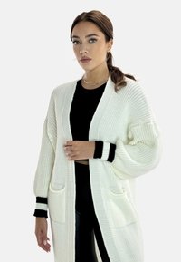 Cardigan bianco lavorato a maglia con spalle basse, polsini a costine e due tasche frontali, caratterizzato da strisce nere decorative sulle maniche.
