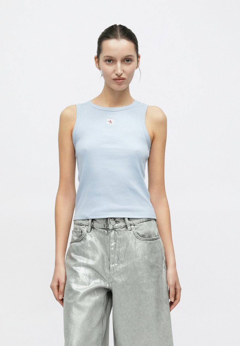 Φωτεινή γαλάζια ριμπ αμάνικη μπλούζα με στρογγυλή λαιμόκοψη και μικρό λογότυπο, συνδυασμένη με λαμπερά ασημένια τζιν παντελόνια φαρδιάς γραμμής.