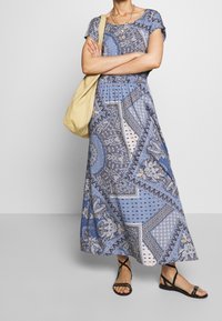 Vestido maxi de tela azul con intrincados patrones de paisley y geométricos, mangas cortas y un corpiño ajustado, combinado con sandalias negras.