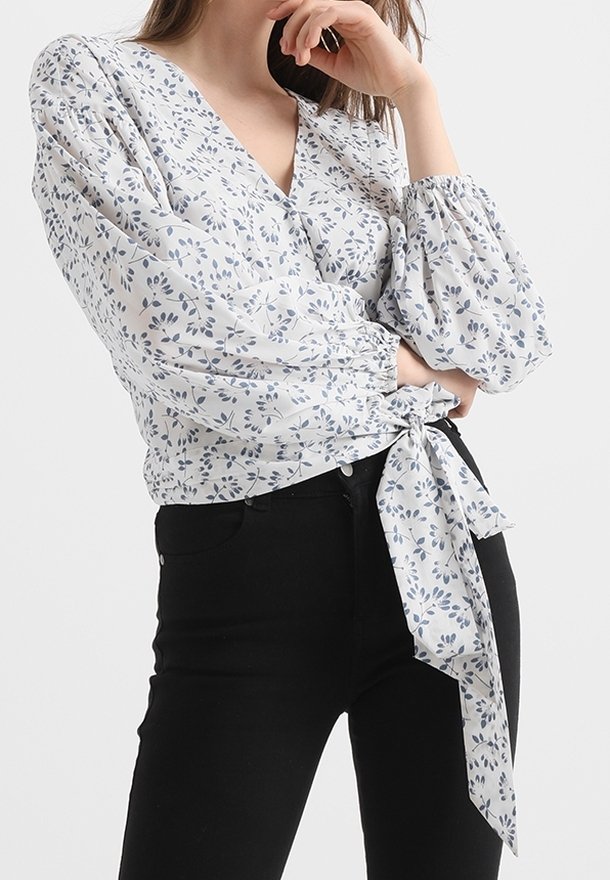 Blouse blanche avec imprimé floral bleu, décolleté en V, manches bouffantes et détail de taille nouée, associée à un pantalon noir ajusté.