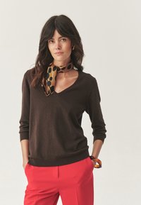 Donna con capelli scuri che indossa un maglione marrone a scollo a V, pantaloni rossi, una sciarpa a motivi leopardati e un braccialetto color ambra, in posa con le mani in tasca.