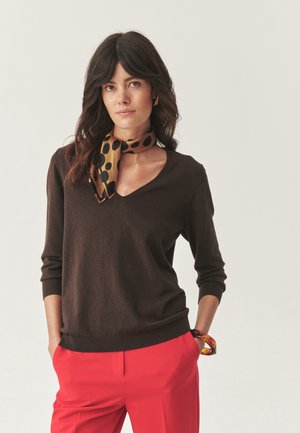 Donna con capelli scuri che indossa un maglione marrone a scollo a V, pantaloni rossi, una sciarpa a motivi leopardati e un braccialetto color ambra, in posa con le mani in tasca.