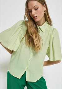 Camicia verde chiaro con bottoni, maniche corte e voluminose, e colletto a punta. Il tessuto presenta un aspetto testurizzato. Abbinata a pantaloni verdi.