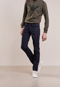 Tummansiniset farkut, joissa on slim fit -leikkaus, yhdistettynä vihreään collegepaitaan, jossa on logo. Asun viimeistelee valkoiset kudotut slip-on -kengät.