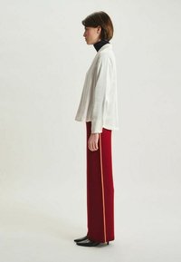 Haut blanc à manches longues avec une coupe décontractée, associé à un pantalon rouge taille haute avec une bande orange sur le côté et des chaussures noires élégantes.