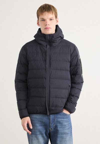 Doudoune bleu marine avec un col haut et une fermeture éclair. Elle présente un motif matelassé horizontal et une patch sur la manche gauche.