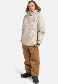 Mężczyzna z kręconymi włosami, ubrany w jasną, wzorzystą kurtkę z kapturem, brązowe spodnie i czarne zimowe buty, stoi z rękami w kieszeniach.