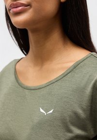 Κοντινό πλάνο στον λαιμό και τους ώμους ενός ατόμου που φοράει πουκάμισο ελαιί πράσινου χρώματος με ένα μικρό λευκό στυλιζαρισμένο λογότυπο στο στήθος.