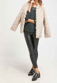 Faux furjacka i ljusbeige, långa ärmar, parat med en mörkgrön blus, svarta läderbyxor och svarta skor med spännen.