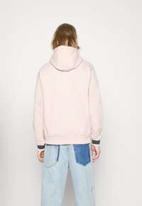 Sudadera de color rosa empolvado con cordón y una textura suave y lisa. Presenta puños y dobladillo acanalados con un borde en contraste. Llevada sobre unos vaqueros de color azul claro.