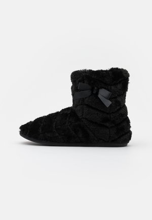 Bottine en fausse fourrure noire avec texture matelassée, présentant un design en volume et un nœud en satin sur le côté. Semelle plate.