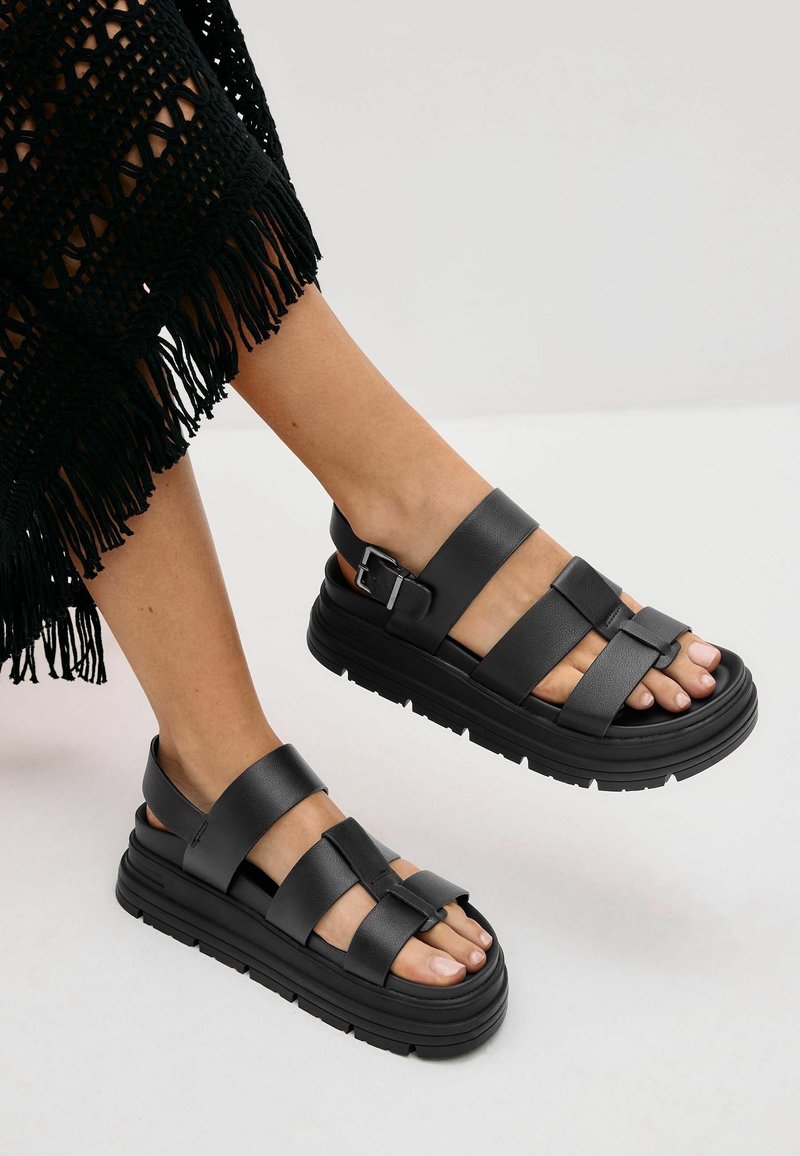 Pieds chaussés de sandales noires à semelles épaisses avec trois larges brides et des boucles à la cheville, assortis à une jupe noire à franges.