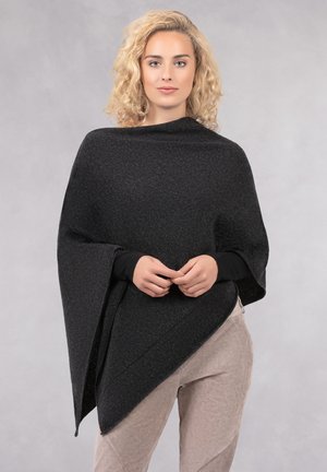 Poncho en laine noire au design drapé, avec un ourlet asymétrique et un col montant, porté sur un haut noir ajusté et un pantalon marron clair.