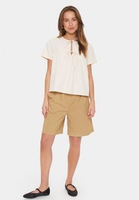 Blusa in cotone beige chiaro con dettaglio a chiave e fiocco, abbinata a pantaloni corti khaki. Il modello indossa delle ballerine nere. Sfondi semplici.