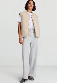 Gilet trapuntato beige sopra una maglietta bianca, abbinato a pantaloni della tuta grigi a gamba larga. Scarpe slip-on nere con motivi circolari bianchi.