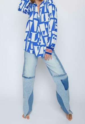 Osoba nosząca oversize'ową niebiesko-białą koszulę z długim rękawem z nadrukiem, w połączeniu z luźnymi patchworkowymi dżinsami, stojąca boso na jednolitym tle.